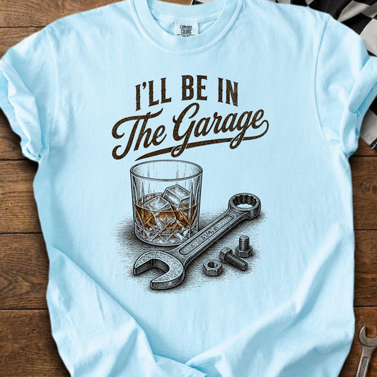 Light blue t-shirt with text "I'LL BE IN The Garage", glass of whiskey, wrench, and nuts.