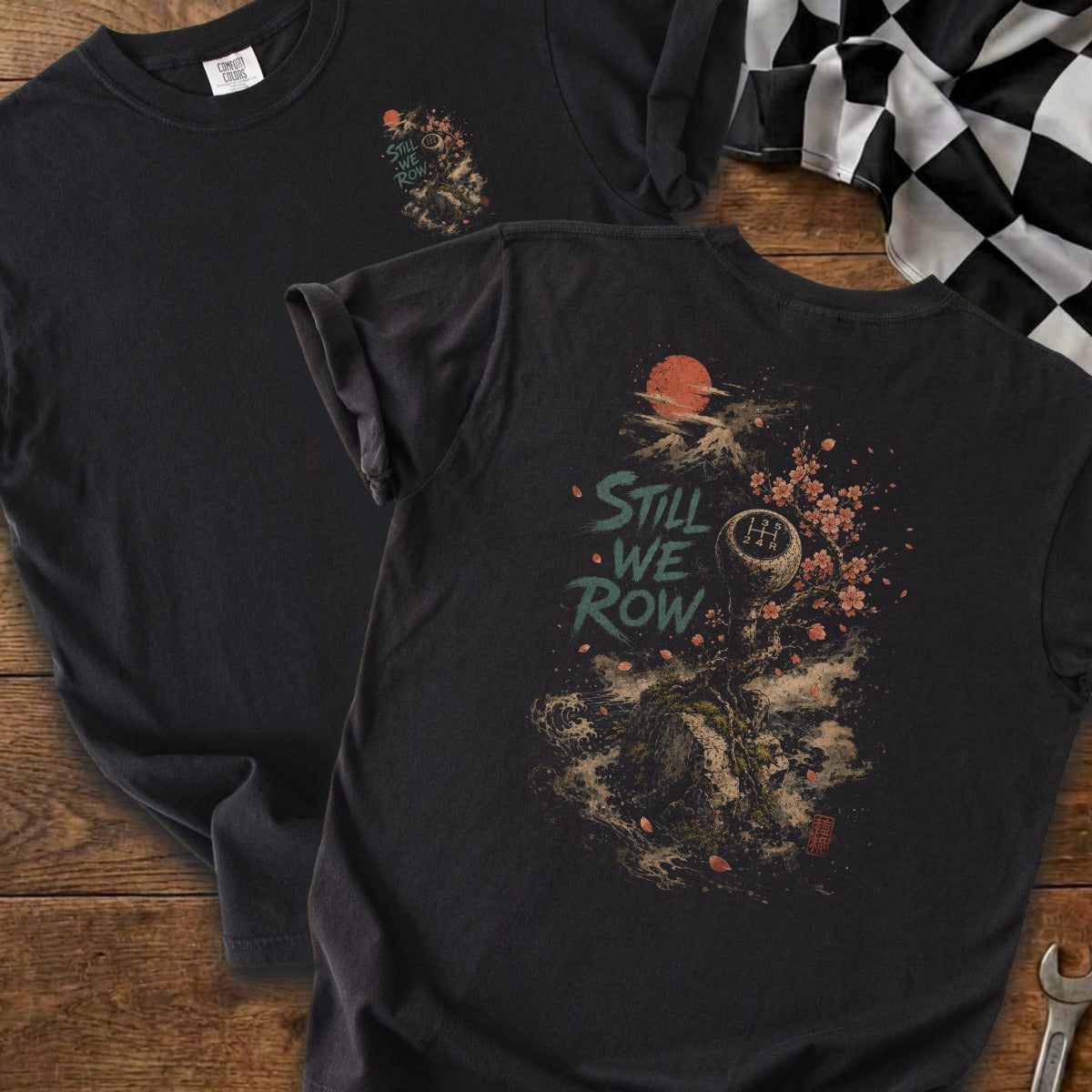 Black t-shirt with graphic art featuring a gear shift, cherry blossoms, sun, and mountain titled "Still We Row".