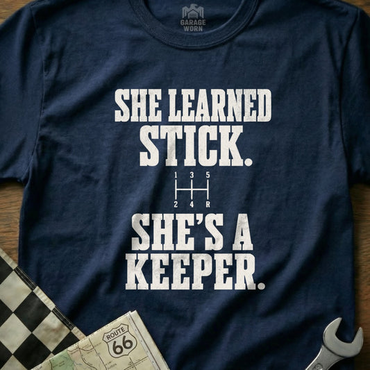 Navy t-shirt with white text reading "SHE LEARNED STICK. SHE'S A KEEPER."