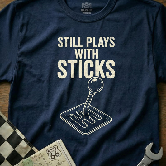 Navy t-shirt with "Still Plays with Sticks" text and a manual gear shift graphic.