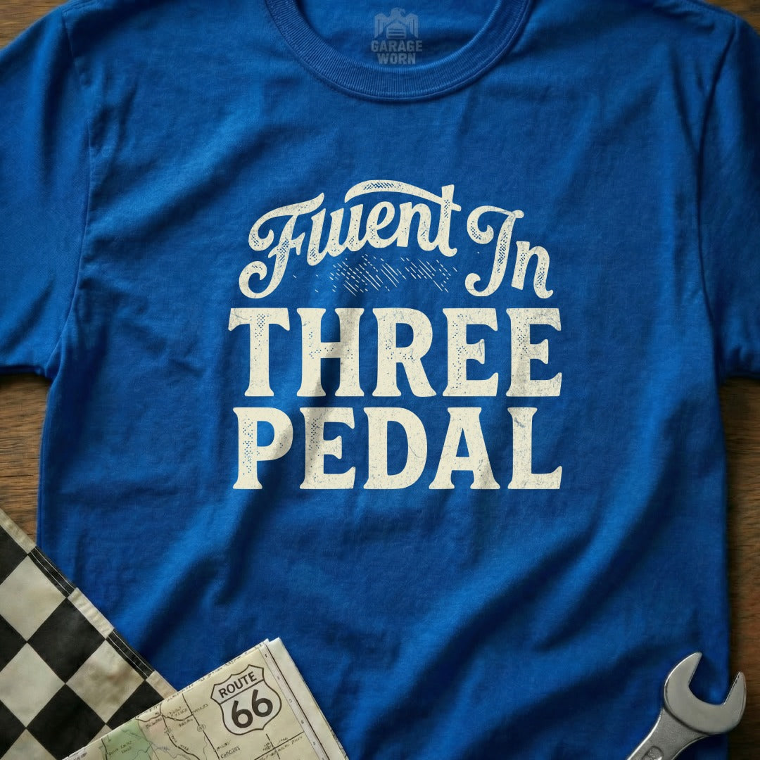 Blue t-shirt with white text "Fluent in THREE PEDAL" and checkered flag detail.
