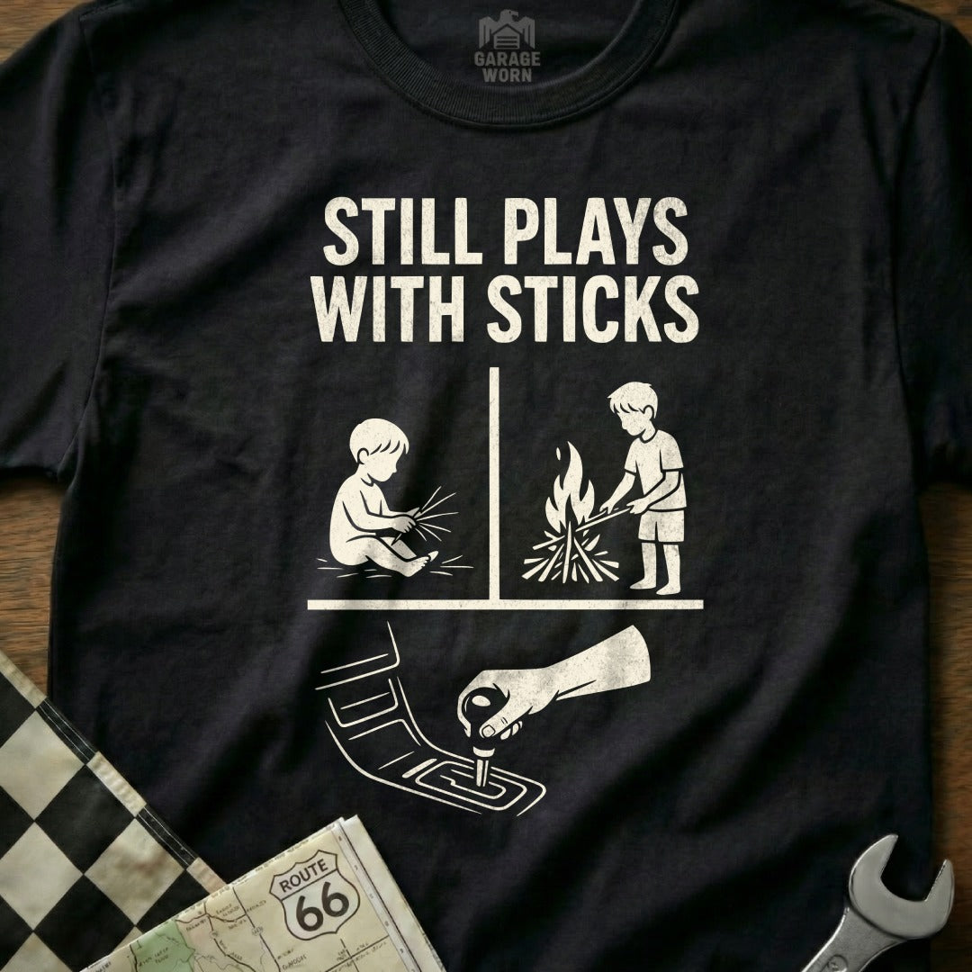 T-shirt with "Still Plays with Sticks" text and graphics of child with fire and child with gear shift.
