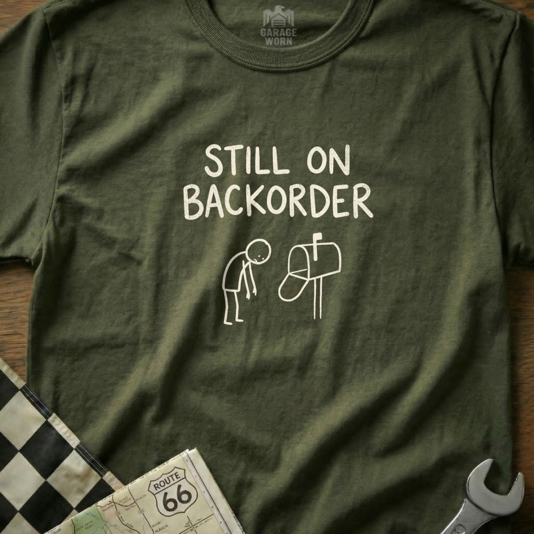 Olive green t-shirt with "Still on Backorder" text and stick figure by mailbox.