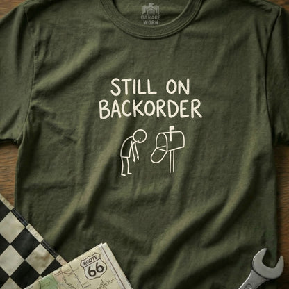 Olive green t-shirt with "Still on Backorder" text and stick figure by mailbox.