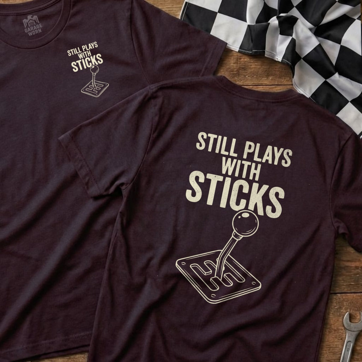Burgundy t-shirt back with graphic of a vintage manual gear shift and text "Still Plays With Sticks".