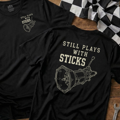 Black t-shirt with "STILL PLAYS WITH STICKS" and a transmission graphic.