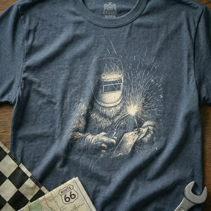 T-shirt featuring a Bigfoot welding with sparks flying, Route 66 map, and checkered flag.