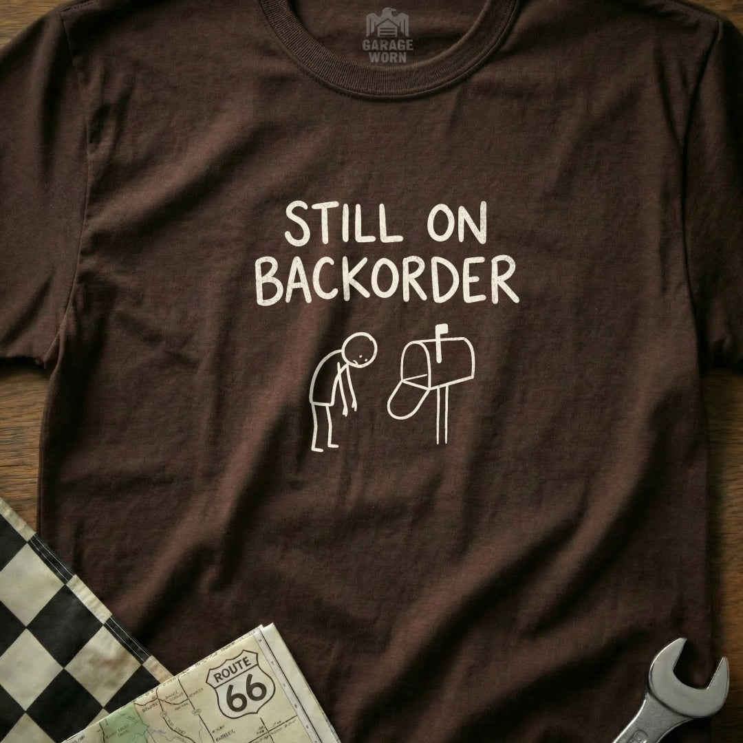 Brown t-shirt with "Still on backorder" text and cartoon of person at mailbox.