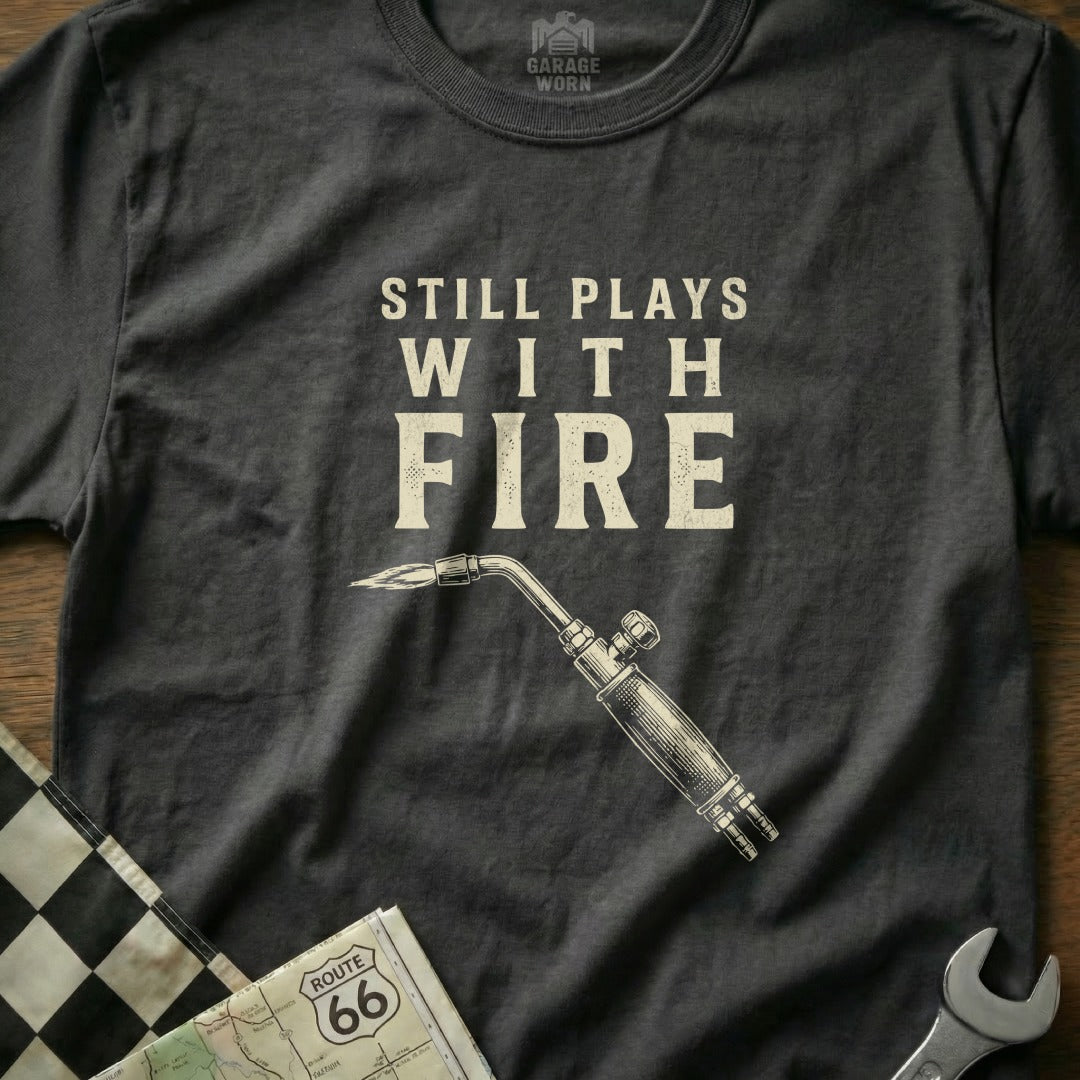 Dark t-shirt with "Still Plays With Fire" text and a welding torch graphic.