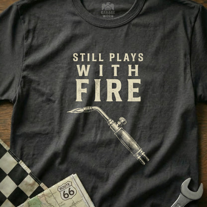 Dark t-shirt with "Still Plays With Fire" text and a welding torch graphic.
