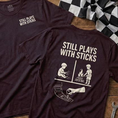 Maroon t-shirt with "Still Plays With Sticks" and controller/campfire graphics.