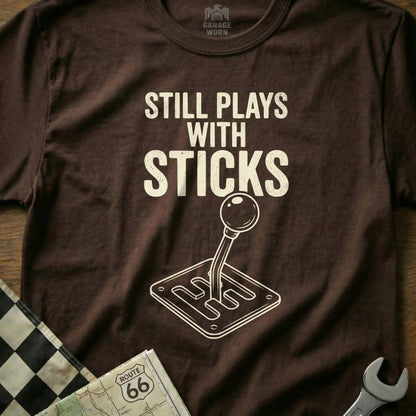 Brown t-shirt with "Still Plays with Sticks" and a manual transmission shift pattern.