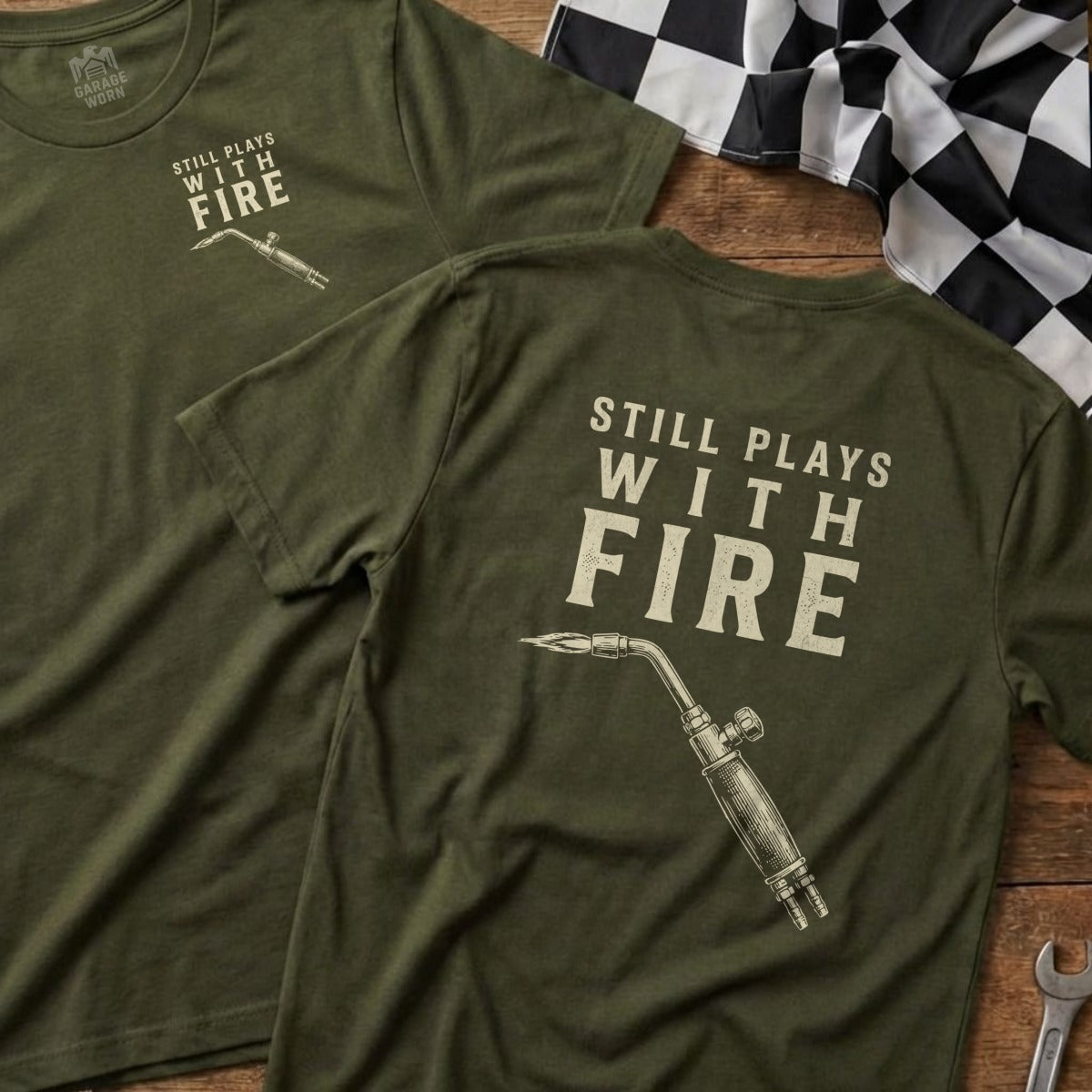 Olive green t-shirt with "Still Plays With Fire" and welding torch graphic.