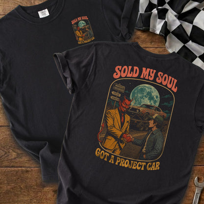 Dark t-shirt graphic of devil shaking hands with man, with car and moon.