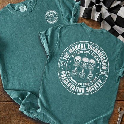 Teal t-shirt with graphic of three skulls with pedals and text "Manual Transmission Preservation Society".