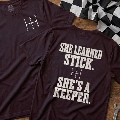 Two maroon t-shirts displaying a manual gear shift pattern and text "She Learned Stick. She's a Keeper."