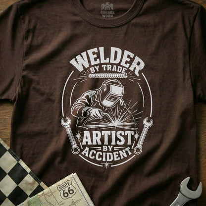 Brown t-shirt with graphic of welder, wrenches, and text "Welder by Trade, Artist by Accident".