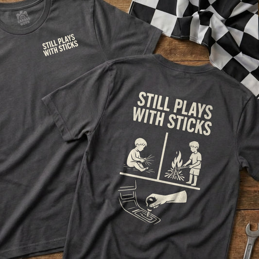 Dark grey t-shirt with "Still Plays With Sticks" graphic of child with joystick and campfire.