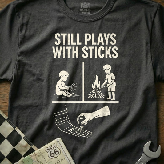 T-shirt with "Still Plays With Sticks" graphic showing baby and child by fire, and car gear shift.