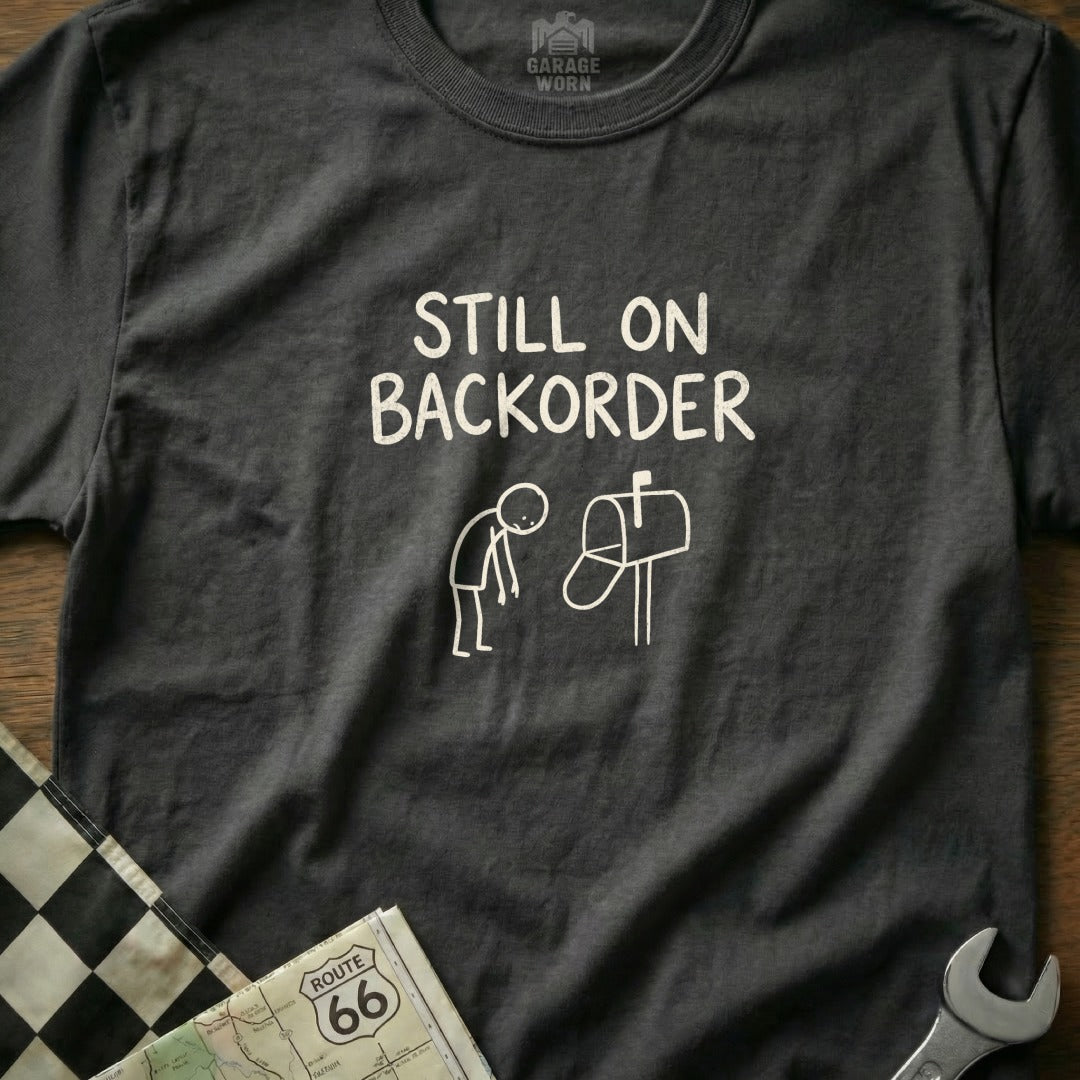 Dark t-shirt with "Still on backorder" text and a stick figure by a mailbox.