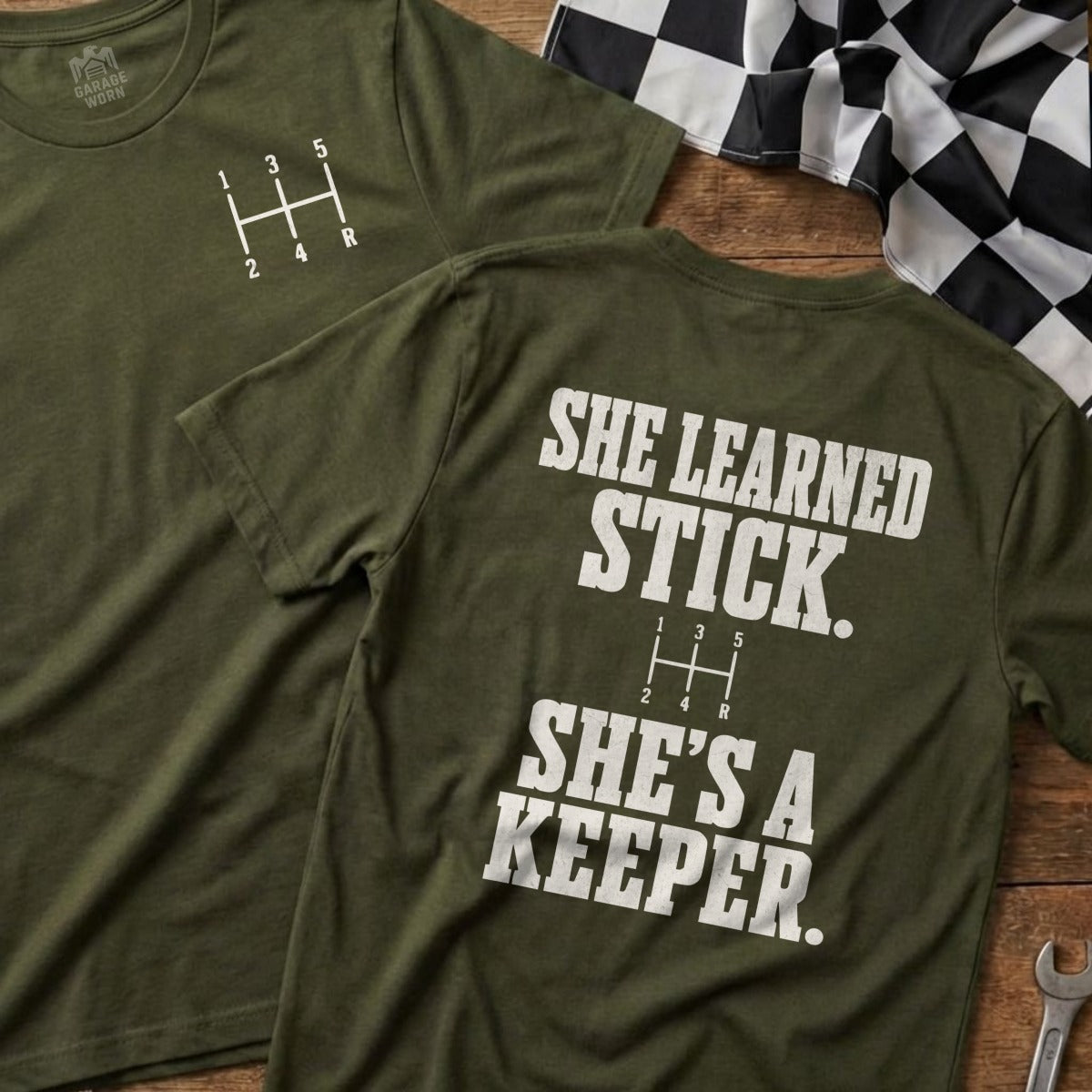 Olive green t-shirt with a manual transmission shift pattern and "SHE LEARNED STICK. SHE'S A KEEPER." text.
