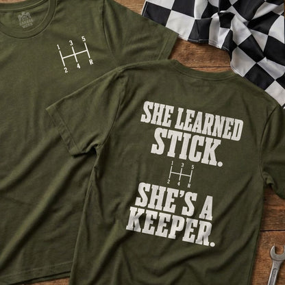 Olive green t-shirt with a manual transmission shift pattern and "SHE LEARNED STICK. SHE'S A KEEPER." text.