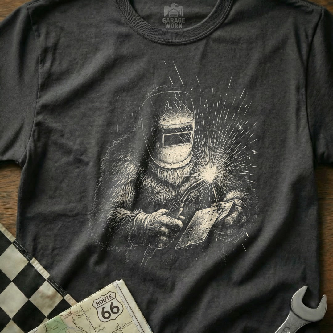 Graphic tee featuring Bigfoot in a welding mask, holding a torch and metal sheet.
