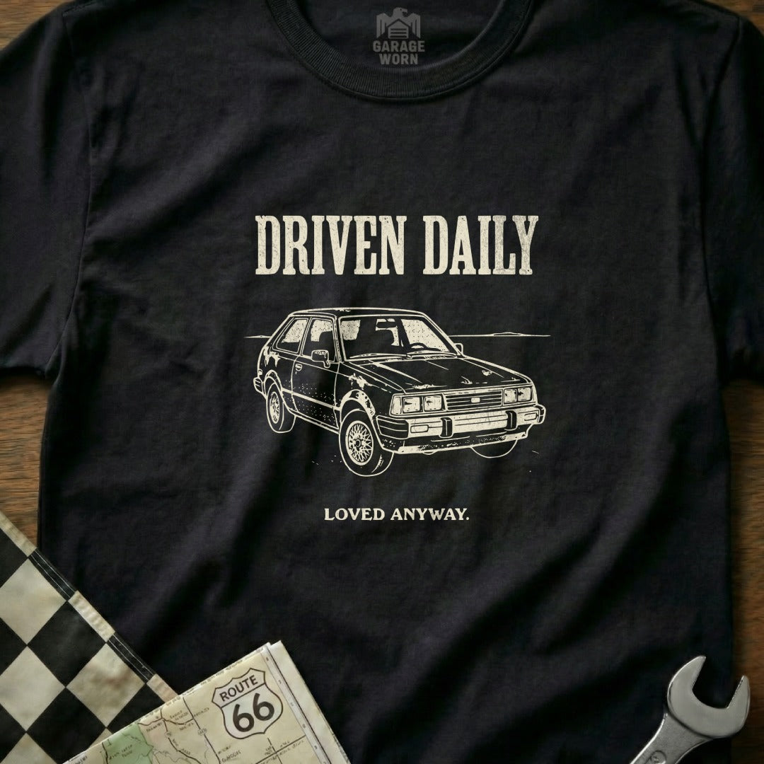 Black t-shirt with white graphic of a vintage car and text "DRIVEN DAILY LOVED ANYWAY."