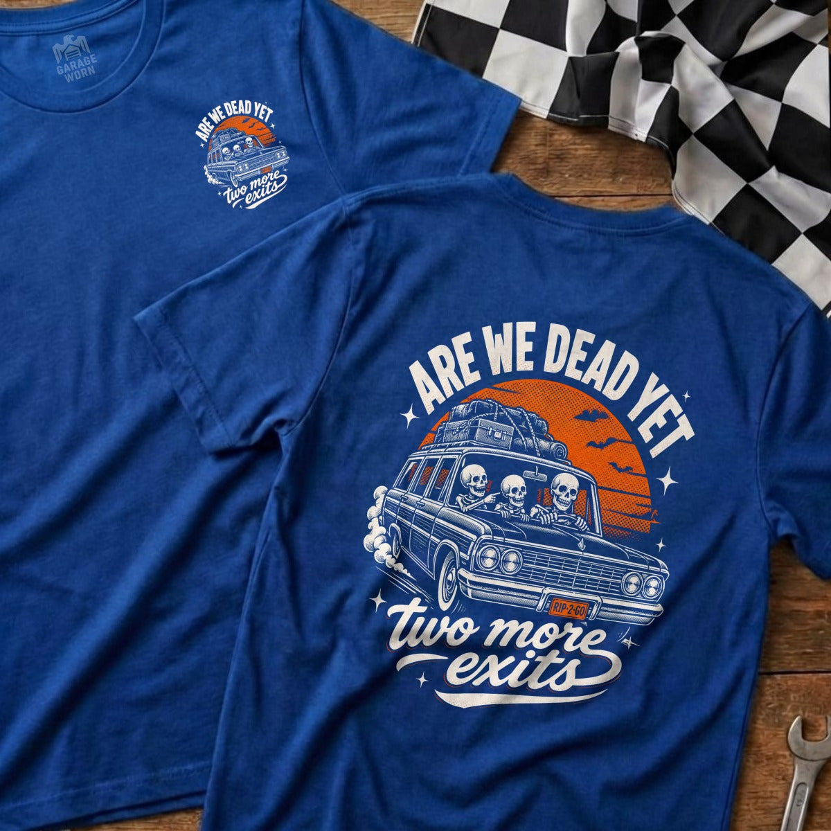 Blue t-shirt with graphic of skeletons driving a car, saying "ARE WE DEAD YET" and "two more exits".