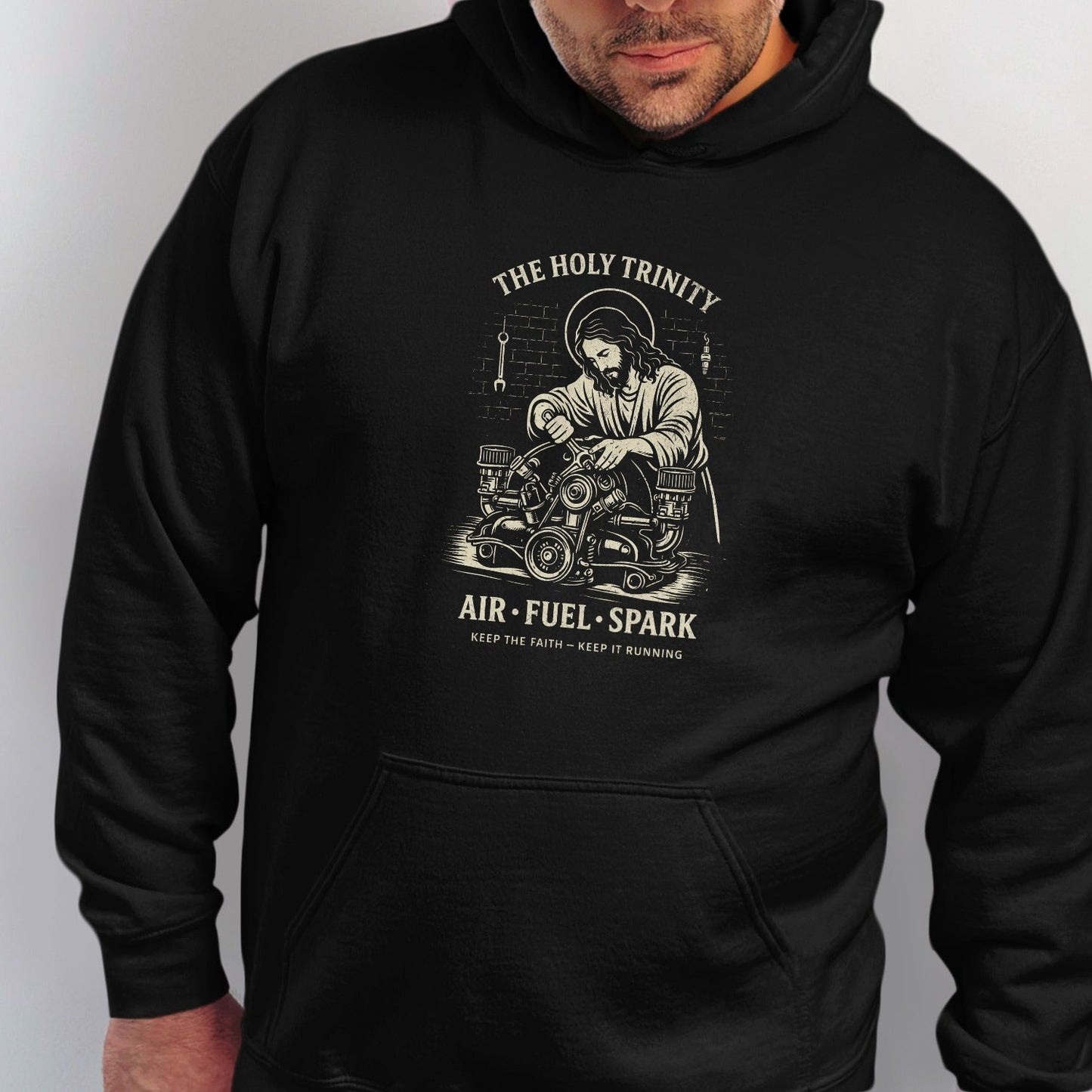 Black hoodie with graphic of Jesus working on an engine, "THE HOLY TRINITY", "AIR FUEL SPARK".