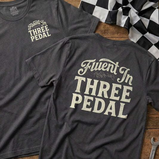 Dark grey t-shirt with "Fluent In Three Pedal" text in off-white.