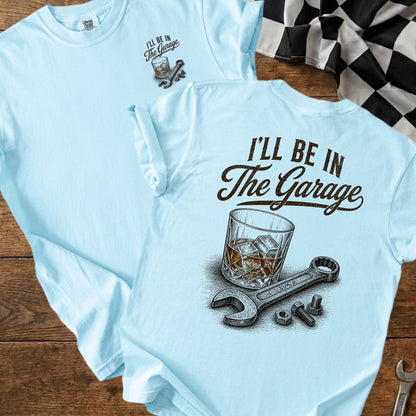 Light blue t-shirt with "I'll Be In The Garage" text, whiskey glass, wrench, nuts, and bolts.