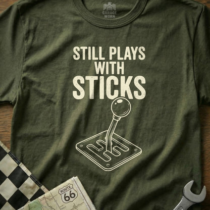 Olive green t-shirt with "Still Plays with Sticks" and manual transmission graphic.