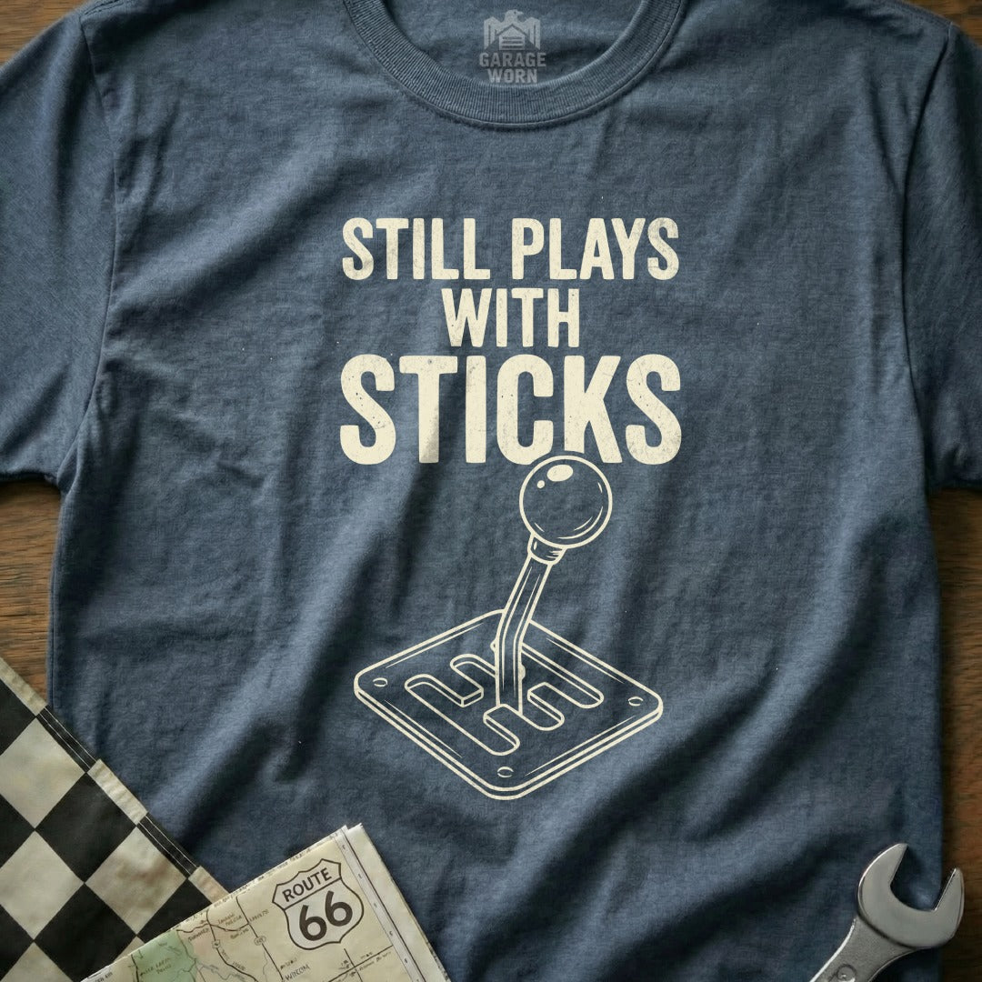 Blue t-shirt with "Still Plays With Sticks" and gear shift graphic.