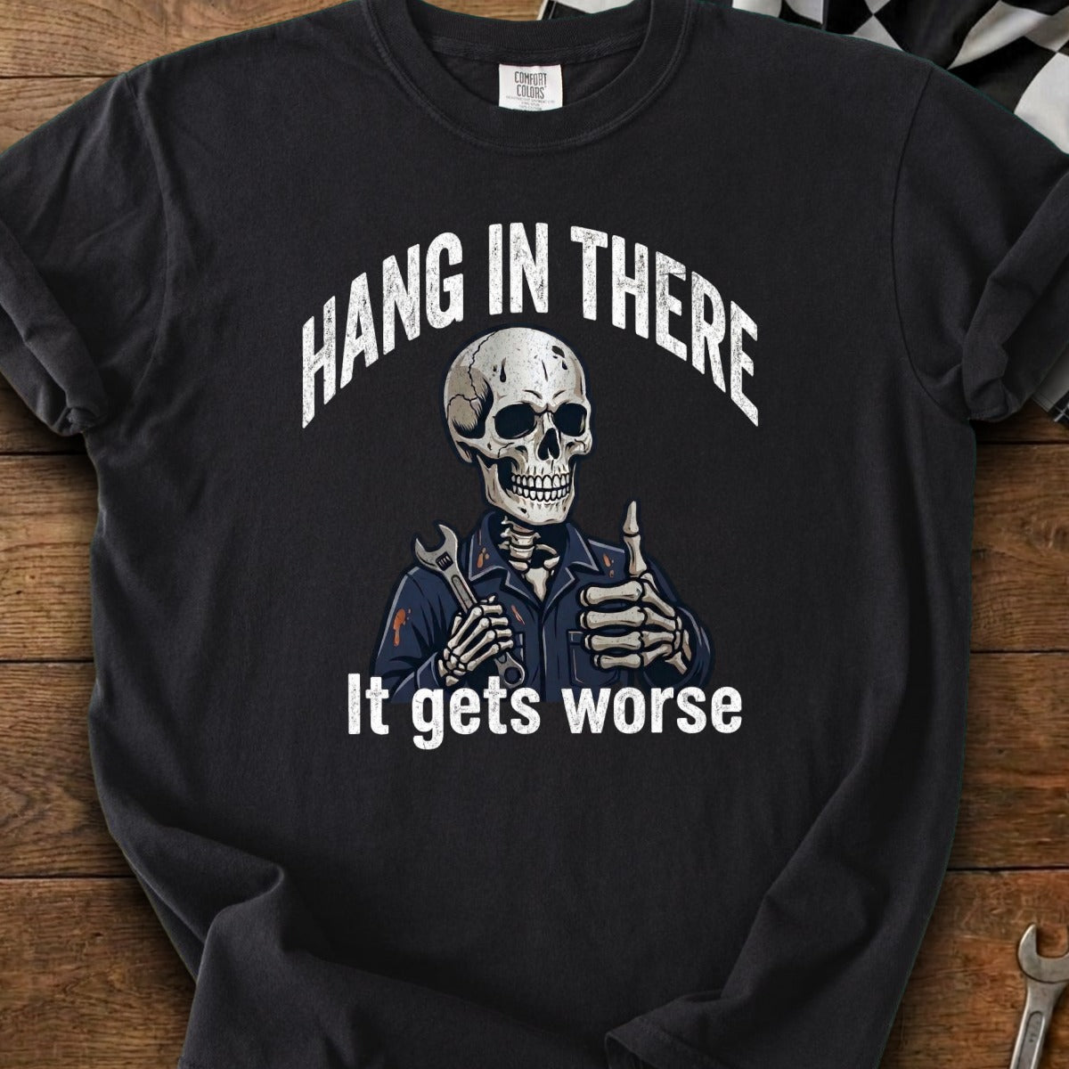 Black t-shirt with skeleton in mechanic's outfit holding a wrench and giving a thumbs up,text "Hang in there. It gets worse."