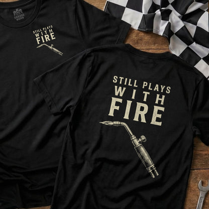 Black t-shirt with "STILL PLAYS WITH FIRE" and a blowtorch graphic.