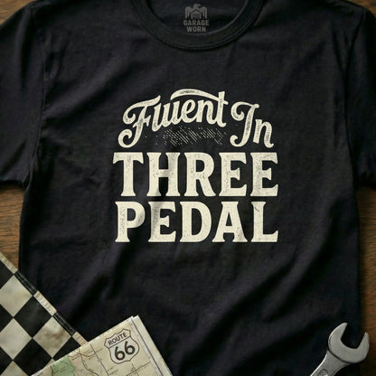 Black t-shirt with "Fluent In THREE PEDAL" in white distressed lettering.