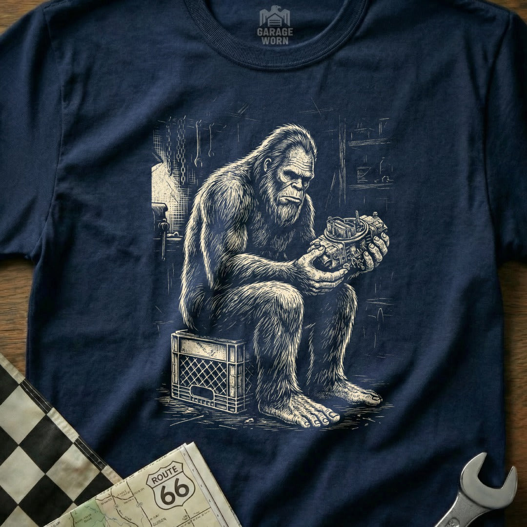 Bigfoot sits on a milk crate holding a carburetor in a workshop.