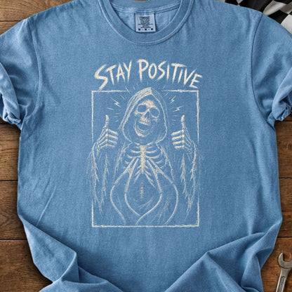Blue t-shirt with a skeleton in a hooded robe giving a thumbs up, with "STAY POSITIVE" text.