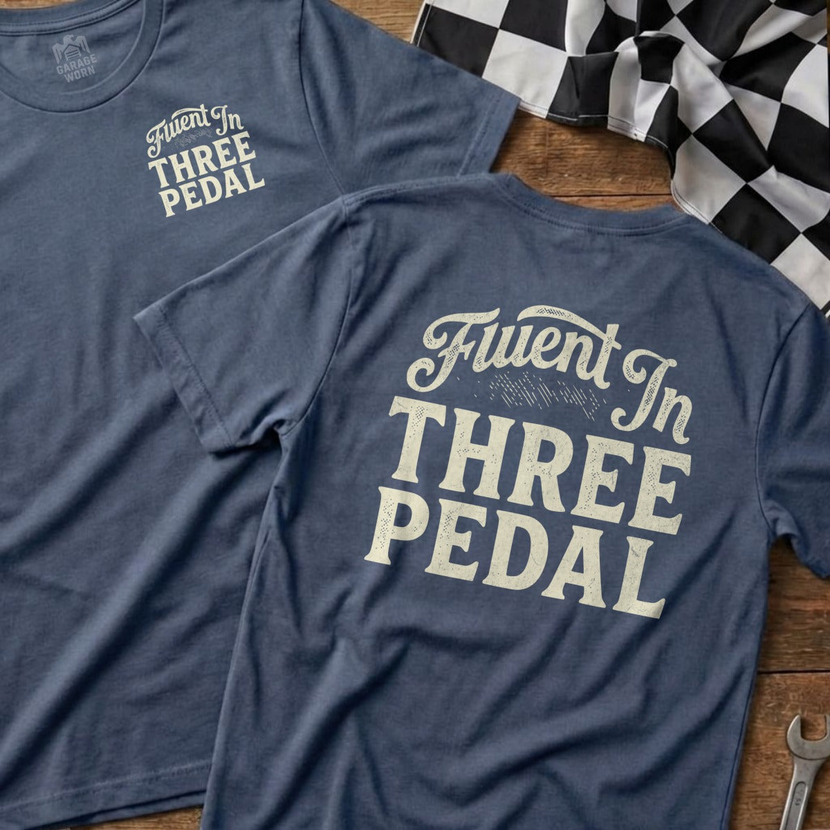 Blue t-shirts with "Fluent In THREE PEDAL" text on the back, on a wooden surface.