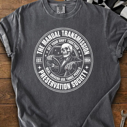 Gray t-shirt with a circular graphic featuring a skeleton driving and text about manual transmissions.