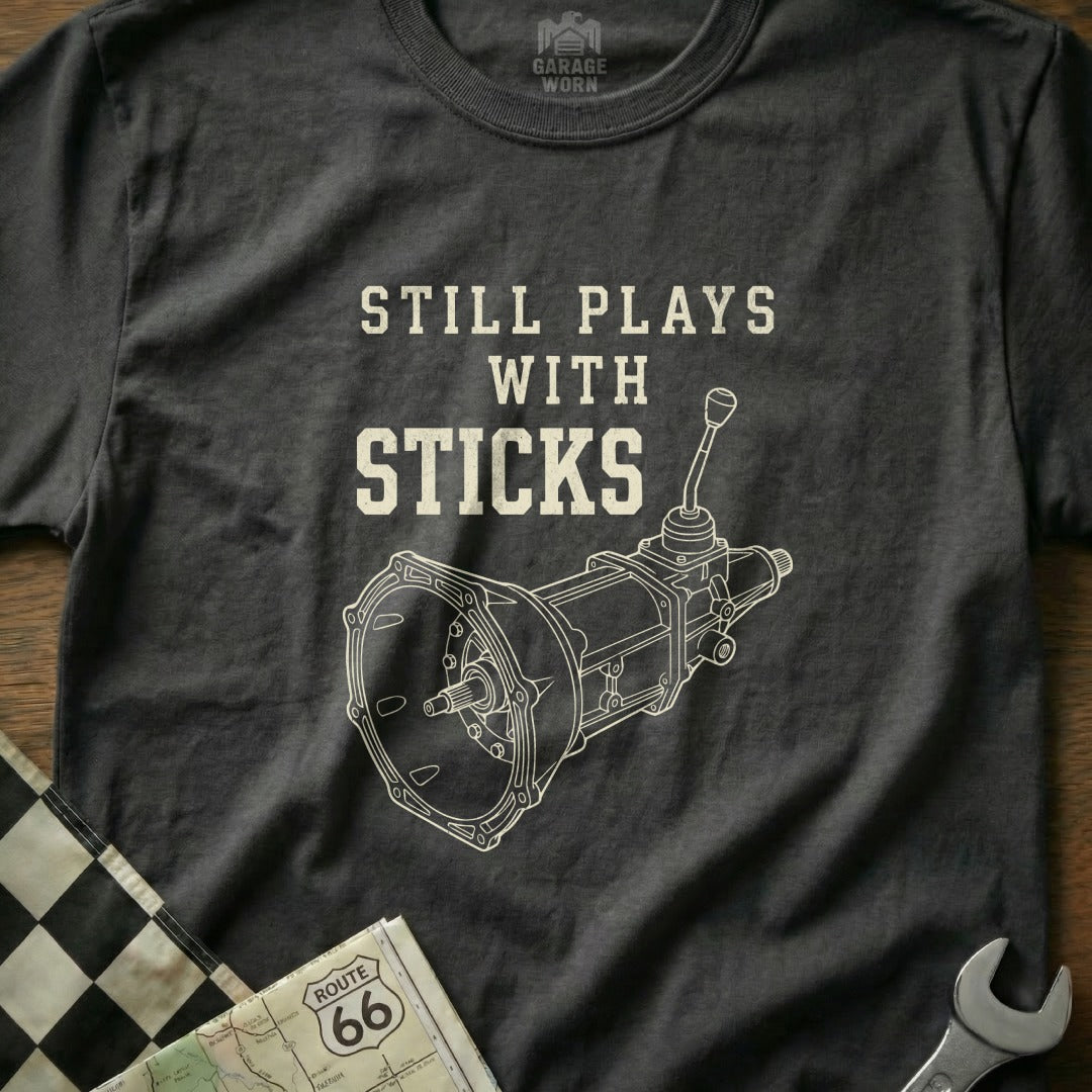Dark gray t-shirt with "STILL PLAYS WITH STICKS" and a manual transmission graphic.