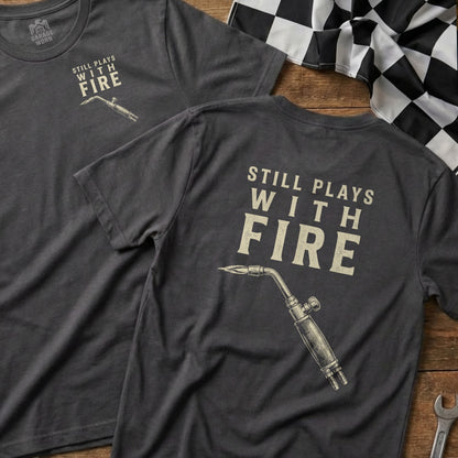 Dark t-shirt with "STILL PLAYS WITH FIRE" and a blow torch graphic.