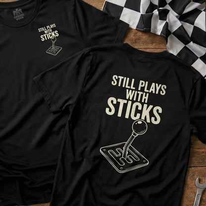 Black t-shirt with "Still Plays With Sticks" and a manual gear shift design.