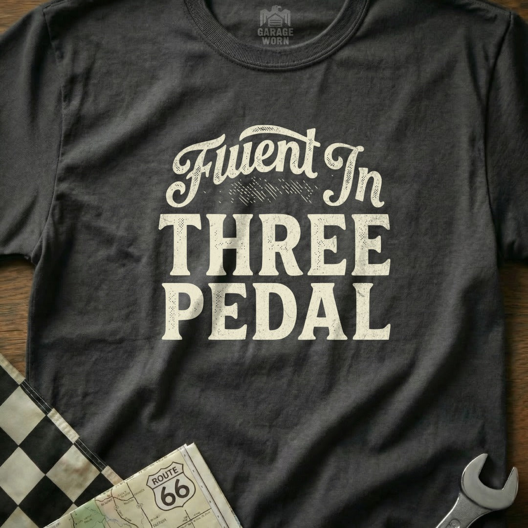 Dark gray t-shirt with "Fluent In Three Pedal" text, checkerboard, and wrench.