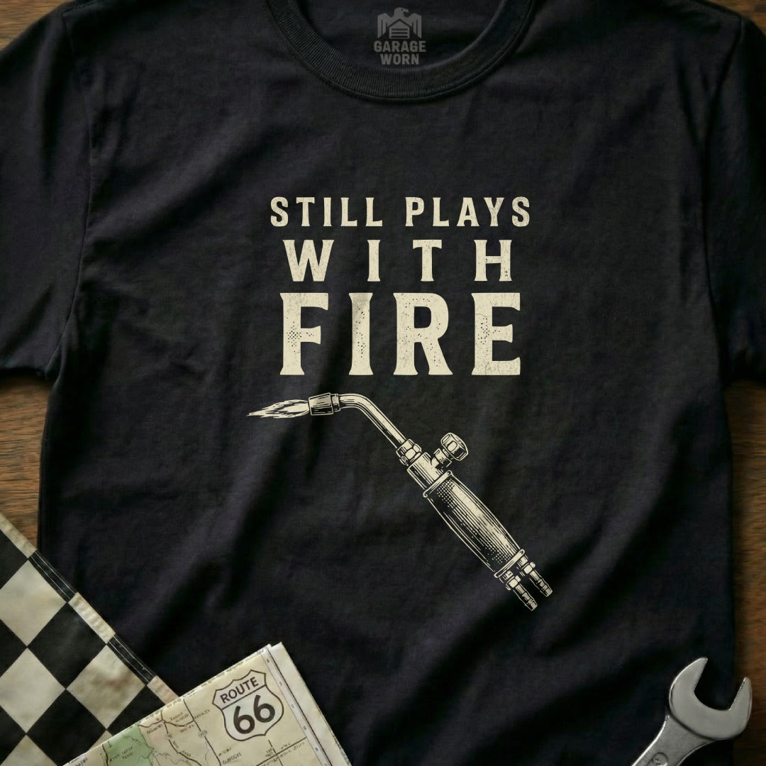 Black t-shirt with "Still Plays With Fire" and a welding torch graphic.