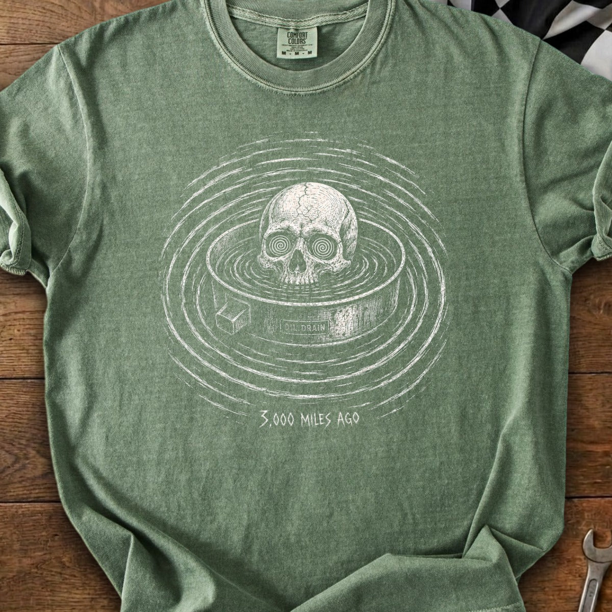 Green t-shirt with skull graphic in a drain labeled "OIL DRAIN" and text "3,000 MILES AGO".