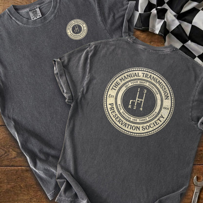 Dark grey t-shirt with circular design featuring a manual gear shift and "The Manual Transmission Preservation Society" text.