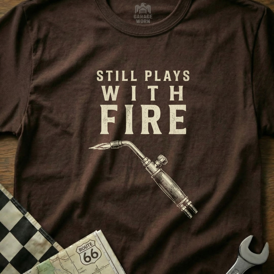 Brown t-shirt with "Still Plays With Fire" and a welding torch.