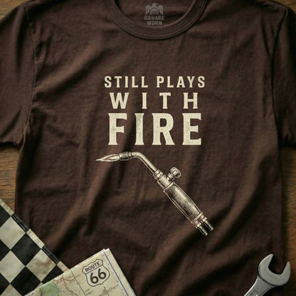 Brown t-shirt with "Still Plays With Fire" and a welding torch.